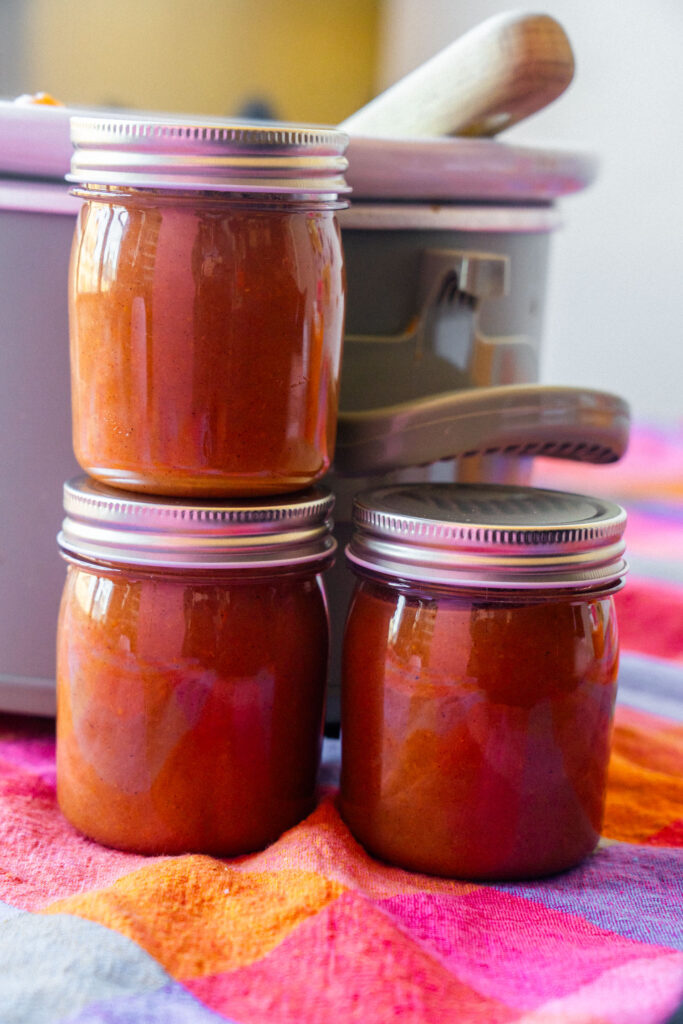 Three glass jars filled with pear butter stacked in front of a slow cooker on a colorful plaid cloth.