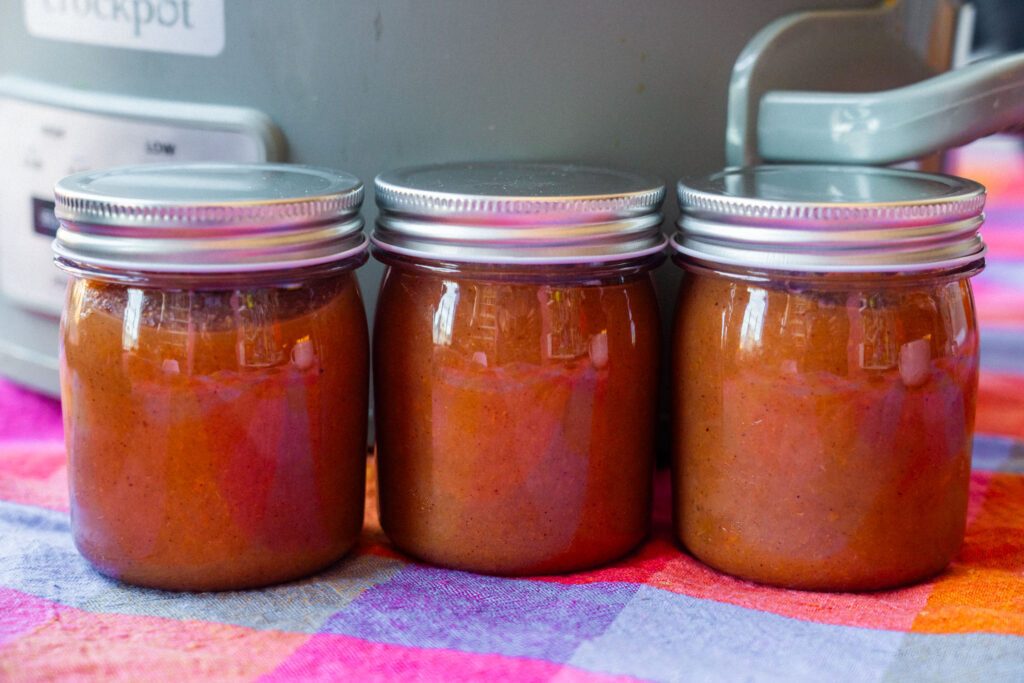 Three small glass jars filled with pear butter, sitting in a row on a plaid cloth in front of a slow cooker.