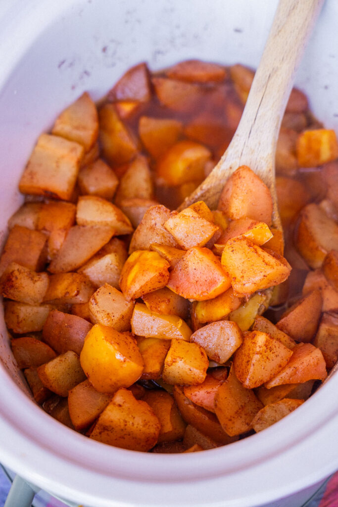 A wooden spoon lifts spiced, cooked pear chunks from a slow cooker.