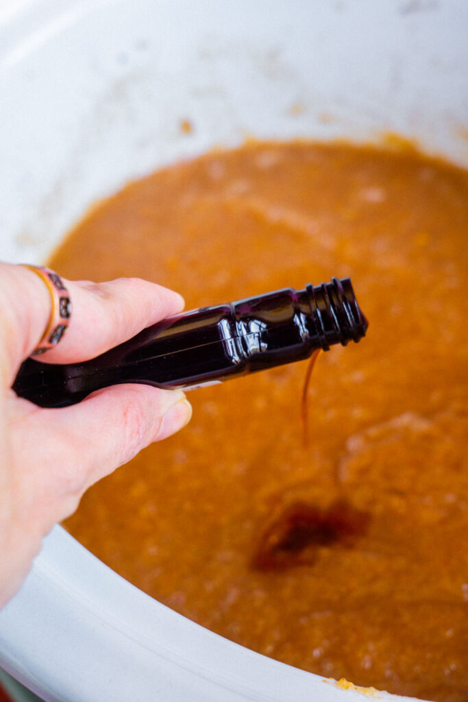 A hand holding a small dark bottle tips a drop of liquid into pear butter in the crockpot.