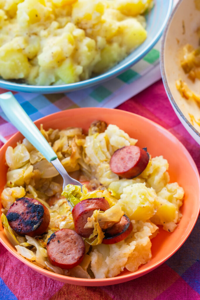 An orange bowl containing a meal of sliced sausage, sautéed cabbage, and potatoes, with a fork holding a piece of sausage.