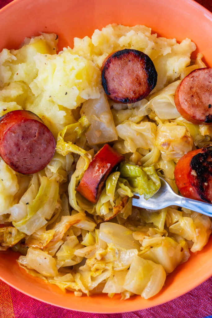 An orange bowl filled with mashed potatoes, cooked cabbage, and sliced browned sausage, with a fork resting in the center.