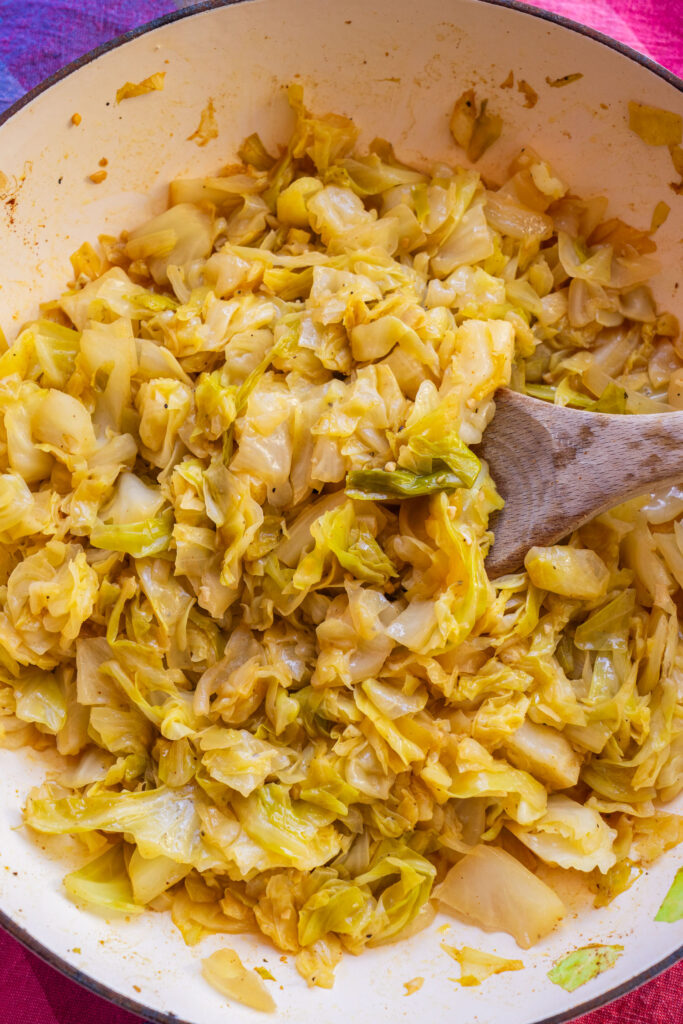 A wooden spoon stirs sautéed chopped cabbage in a light-colored pan.