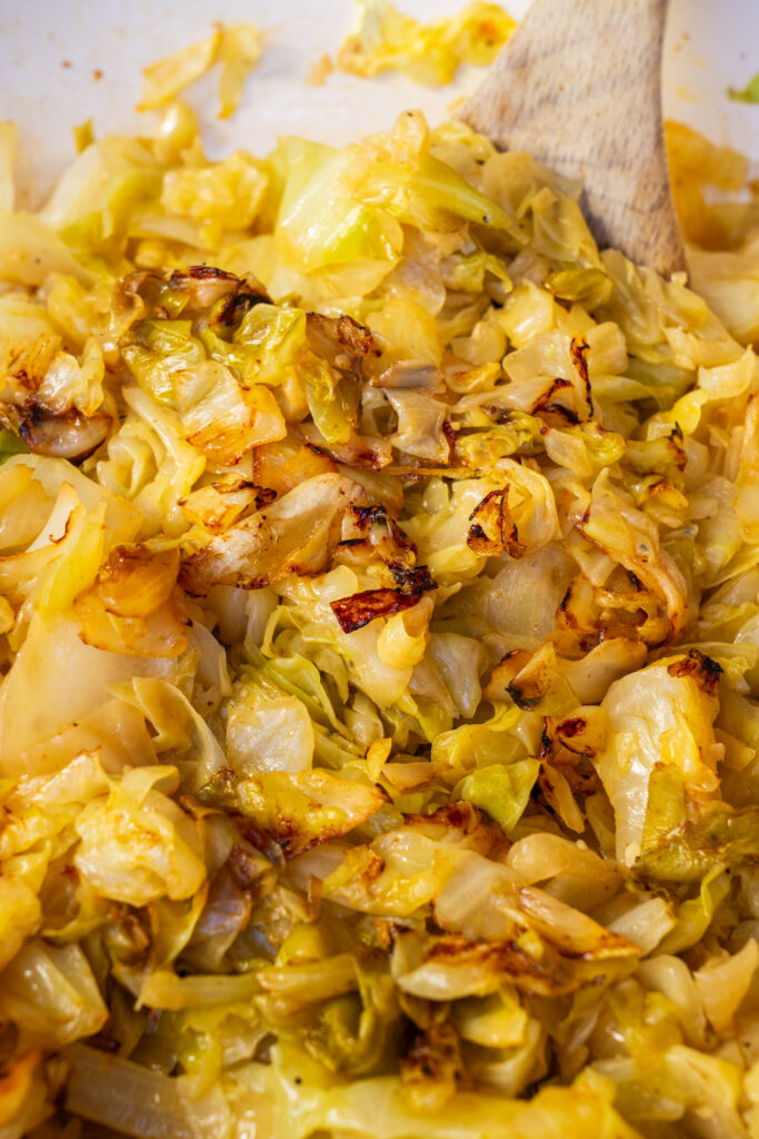 A wooden spoon stirs a pan of sautéed cabbage with browned edges.