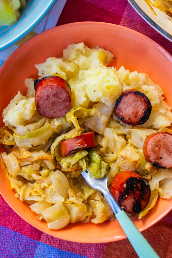 A bowl of mashed potatoes, saut&eacute;ed cabbage, and sliced sausage with a fork.