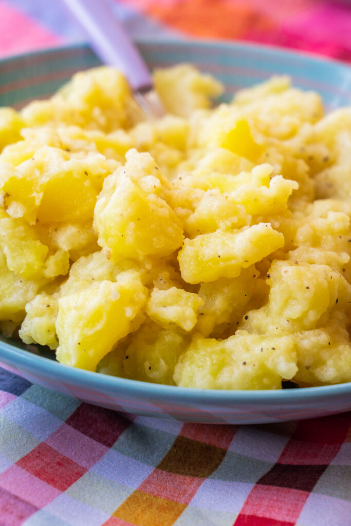 A bowl of seasoned mashed potatoes with a purple spoon resting inside, sitting on a colorful plaid tablecloth.