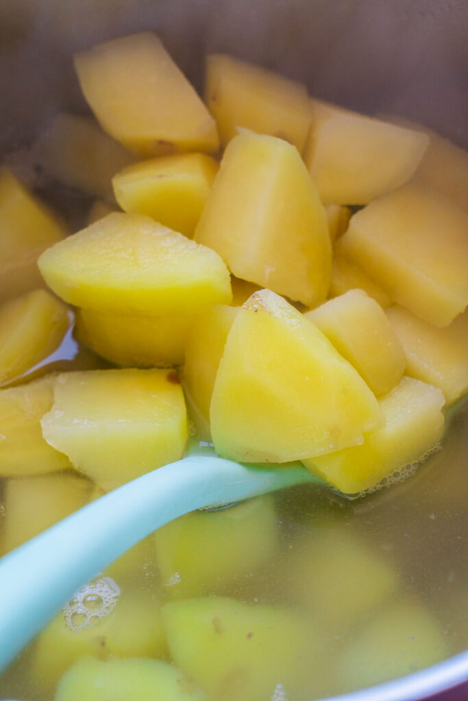 A light blue spoon stirs chunks of boiled potatoes in a pot of water.
