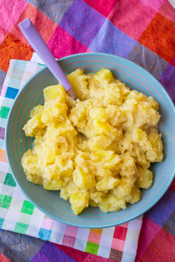 A blue bowl filled with chunky mashed potatoes and a purple spoon, resting on a colorful plaid cloth.